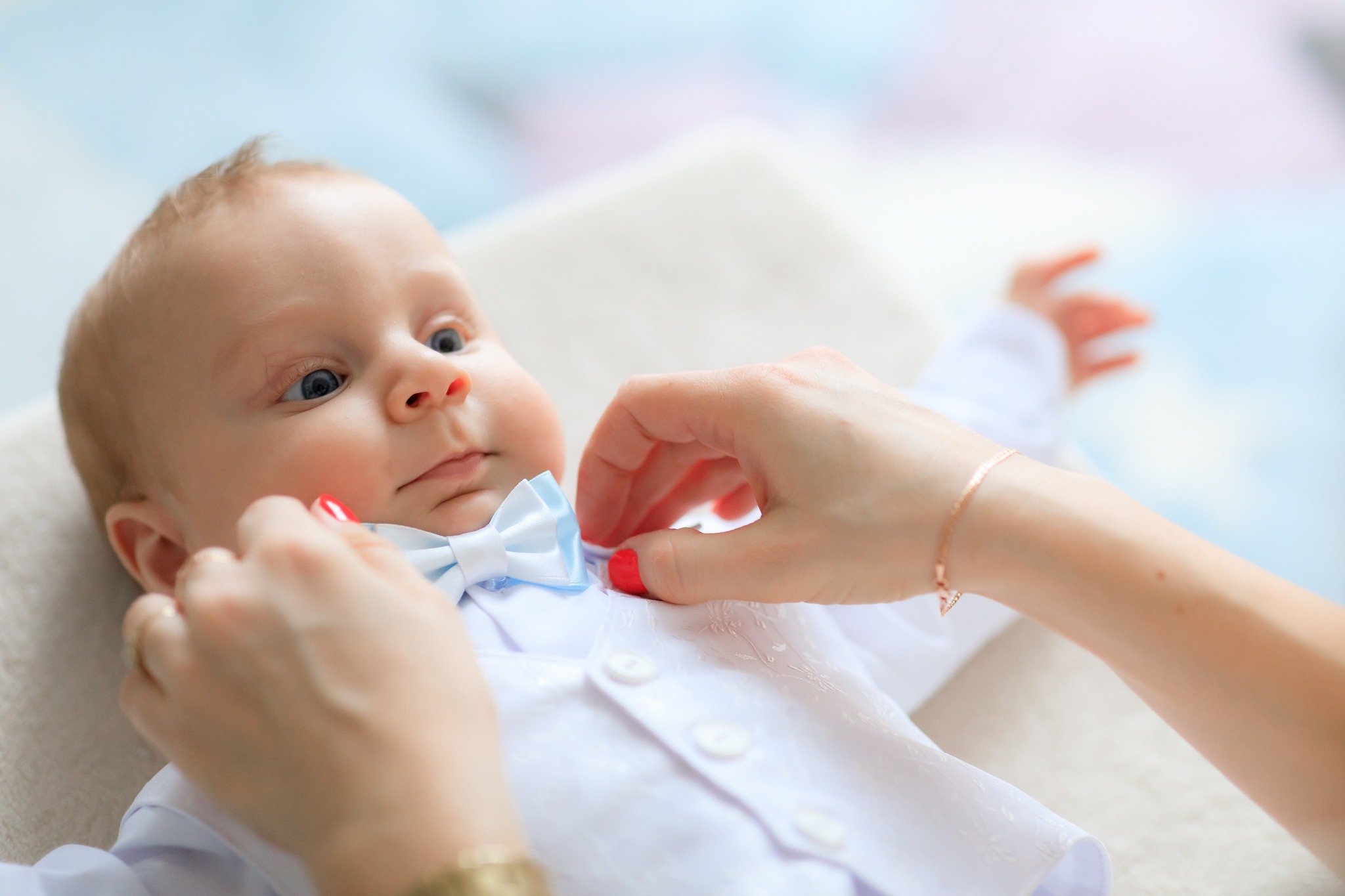 Niemowlę w białej koszuli i błękitnej muszce, dłoń poprawia mu muszkę, jasne tło.
