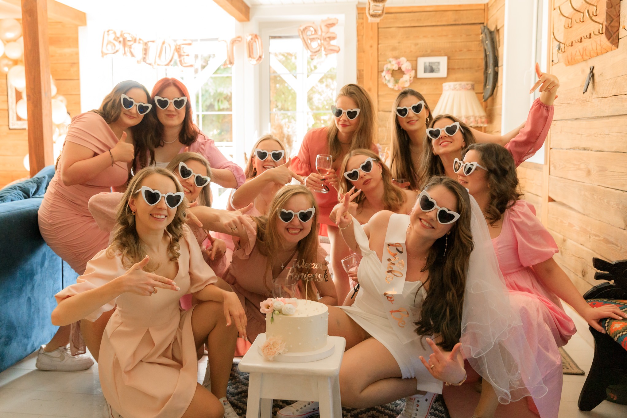 Grupa kobiet w okularach w kształcie serc pozuje do zdjęcia podczas wieczoru panieńskiego w drewnianym wnętrzu, z tortem na pierwszym planie i napisem 'Bride to be' w tle.