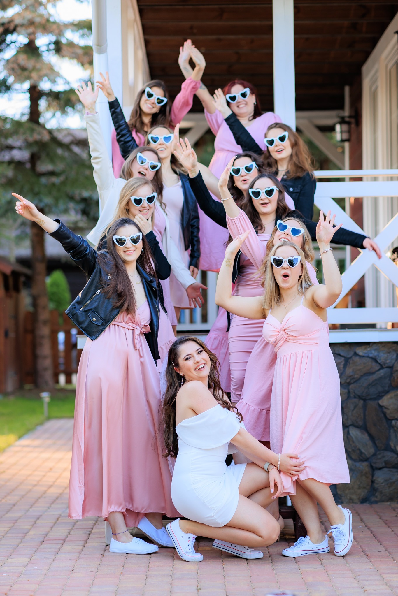 Grupa kobiet w różowych sukienkach i białych okularach w kształcie serc pozuje na zewnątrz, na tle białego budynku z drewnianą balustradą. Jedna kobieta w białej sukience kuca z przodu.