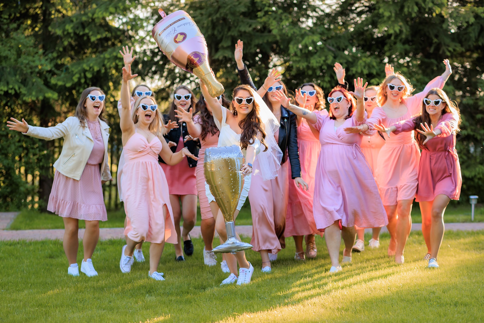 Grupa rozbawionych kobiet w różowych sukienkach i okularach w kształcie serc, świętująca na trawie z balonami w kształcie butelki i kieliszka szampana.