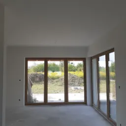 Surowe ściany i podłoga w nowym, jasnym pomieszczeniu z dużymi, drewnianymi oknami wychodzącymi na ogród, widoczne ślady po gładzi.