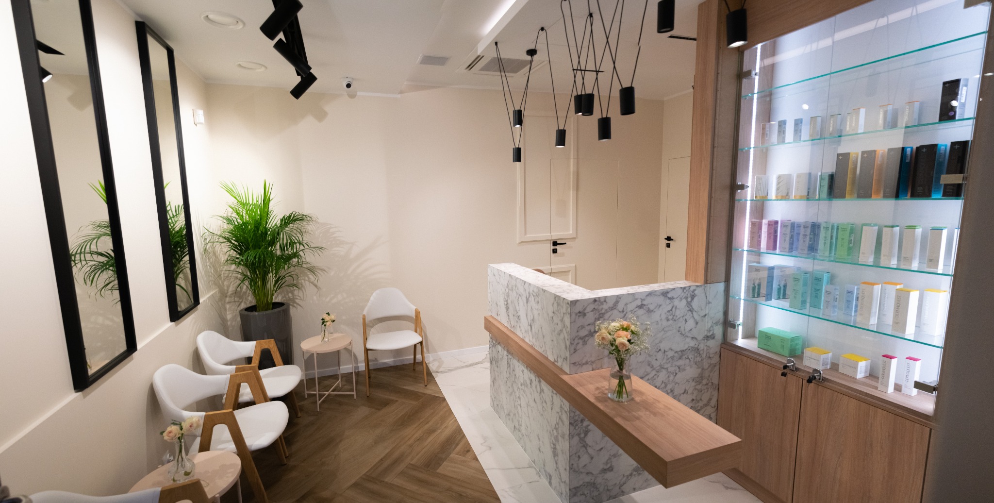 Jasne wnętrze poczekalni: marmurowa lada recepcyjna z kwiatami, rząd krzeseł z drewnianymi nogami, wysoka witryna z produktami kosmetycznymi, podłoga w jodełkę i nowoczesne oświetlenie.