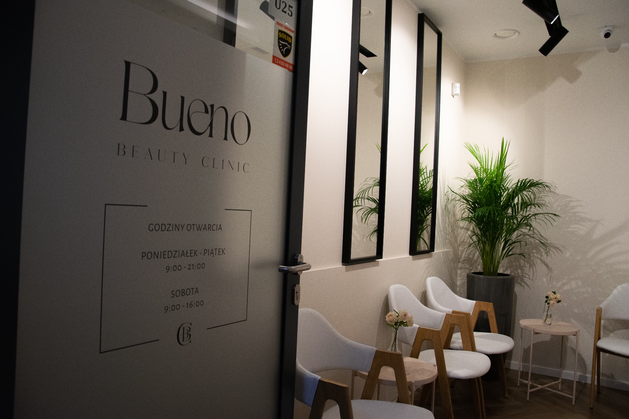 Wnętrze poczekalni salonu kosmetycznego z minimalistycznym wystrojem: białe krzesła z drewnianymi elementami, małe stoliki z kwiatami, duże lustra w czarnych ramach i zielona roślina doniczkowa.