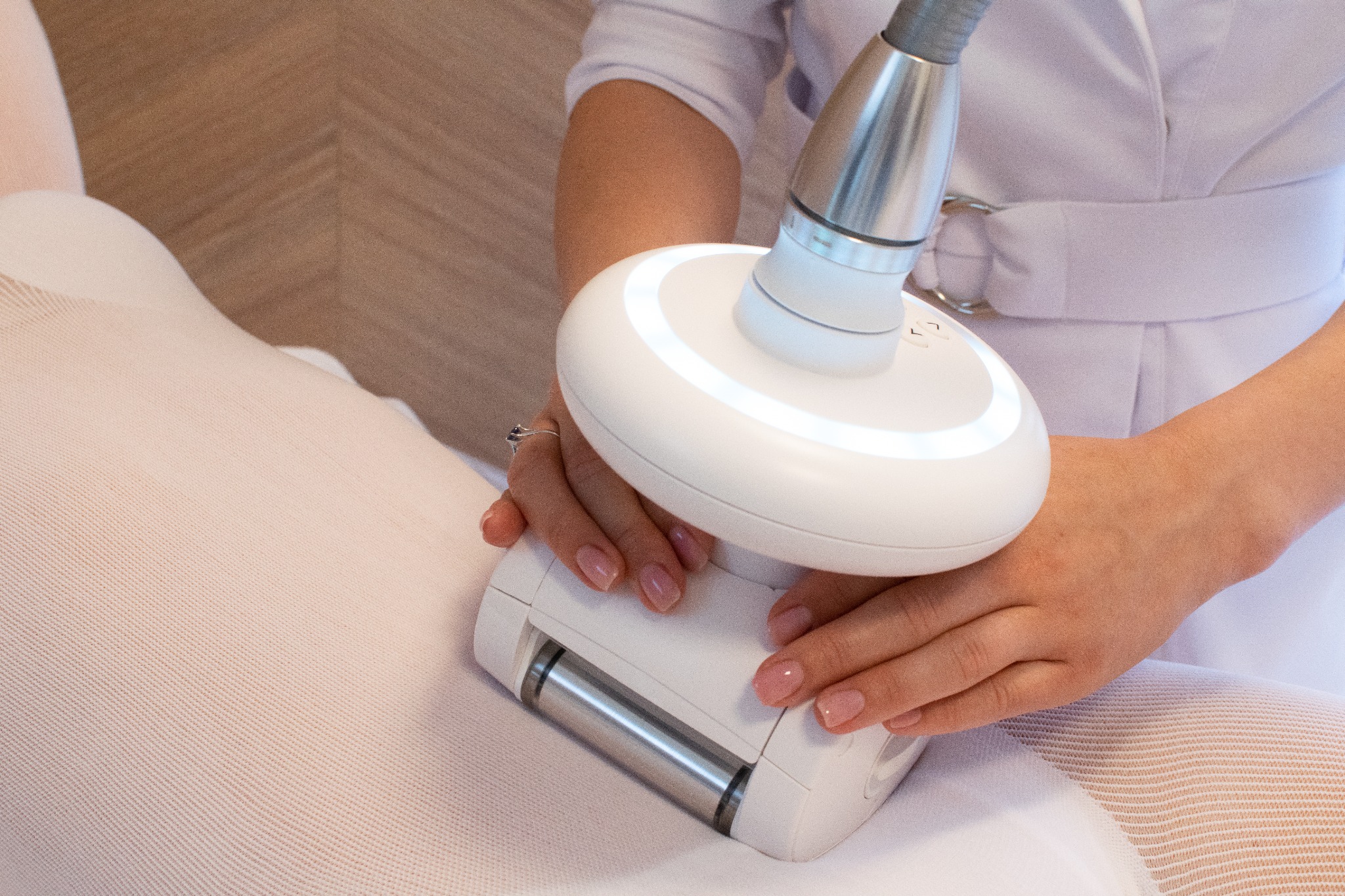 Klientka w kombinezonie podczas zabiegu endermologii LPG, widoczne dłonie kosmetyczki z delikatnym manicure i pierścionkiem oraz głowica urządzenia z podświetleniem LED.