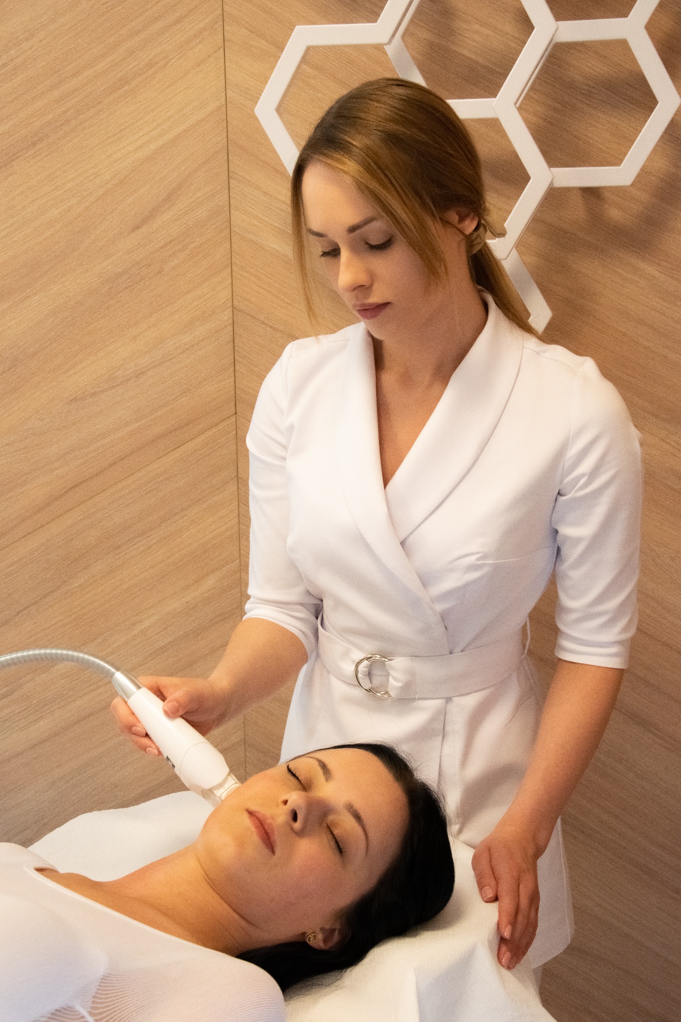 Kobieta w białym uniformie wykonuje zabieg na twarzy klientki leżącej na białym prześcieradle, w tle drewniana ściana z dekoracyjnym białym heksagonem.