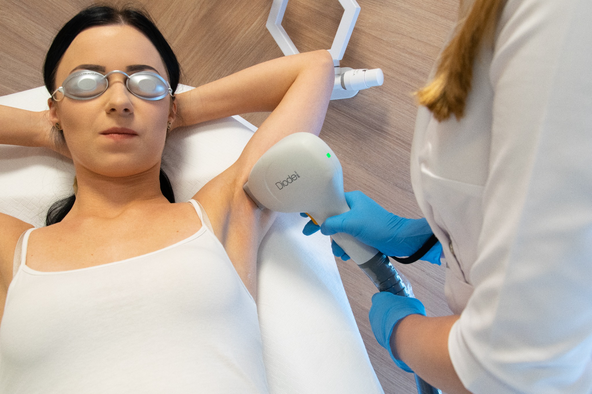 Kobieta w okularach ochronnych leżąca na leżance podczas depilacji laserowej pach. Widoczny laser diodowy w rękach specjalisty w niebieskich rękawiczkach.