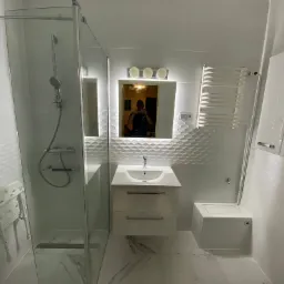 Nowoczesna łazienka z kabiną prysznicową, umywalką z szafką, podświetlanym lustrem i grzejnikiem drabinkowym; białe płytki z delikatnym wzorem na ścianach, marmurowe płytki podłogowe.