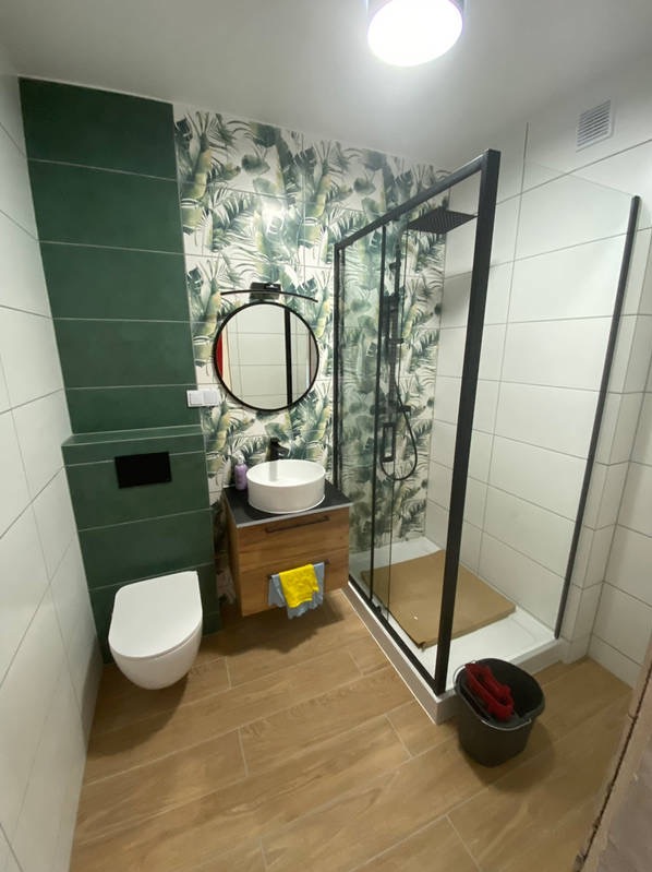 Nowoczesna łazienka z motywem tropikalnym na ścianie, kabina prysznicowa z czarną ramą, okrągłe lustro nad umywalką nablatową, drewniane płytki podłogowe imitujące deski.