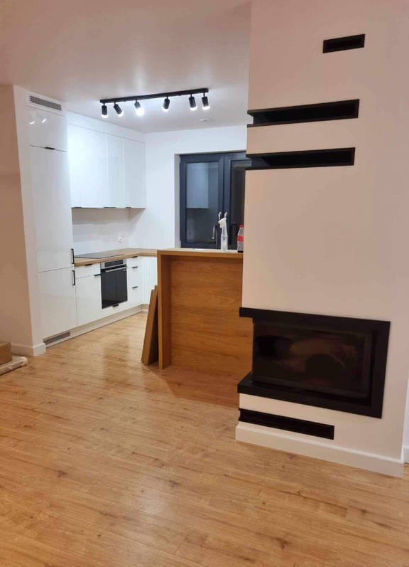 Nowoczesne wnętrze z białymi szafkami kuchennymi, drewnianym blatem, wyspą i kominkiem z czarnymi akcentami, oświetlone reflektorami sufitowymi i położone na podłodze z paneli drewnopodobnych.