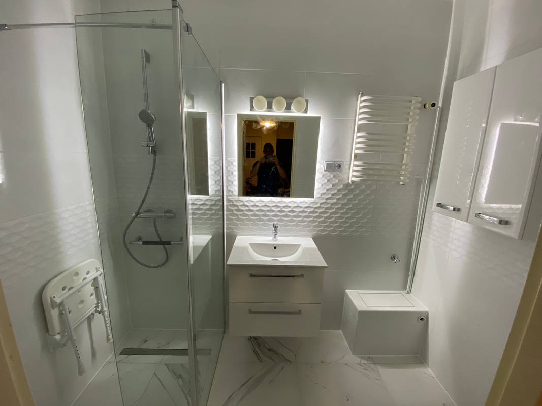 Nowoczesna łazienka z kabiną prysznicową, umywalką z szafką, podświetlanym lustrem i grzejnikiem drabinkowym; białe płytki z delikatnym wzorem na ścianach, marmurowe płytki podłogowe.