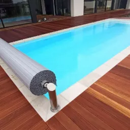 Prostokątny basen z turkusową wodą otoczony drewnianym tarasem z egzotycznego drewna i jasnymi płytami wokół krawędzi basenu, częściowo zwinięta szara roleta na metalowym słupku.