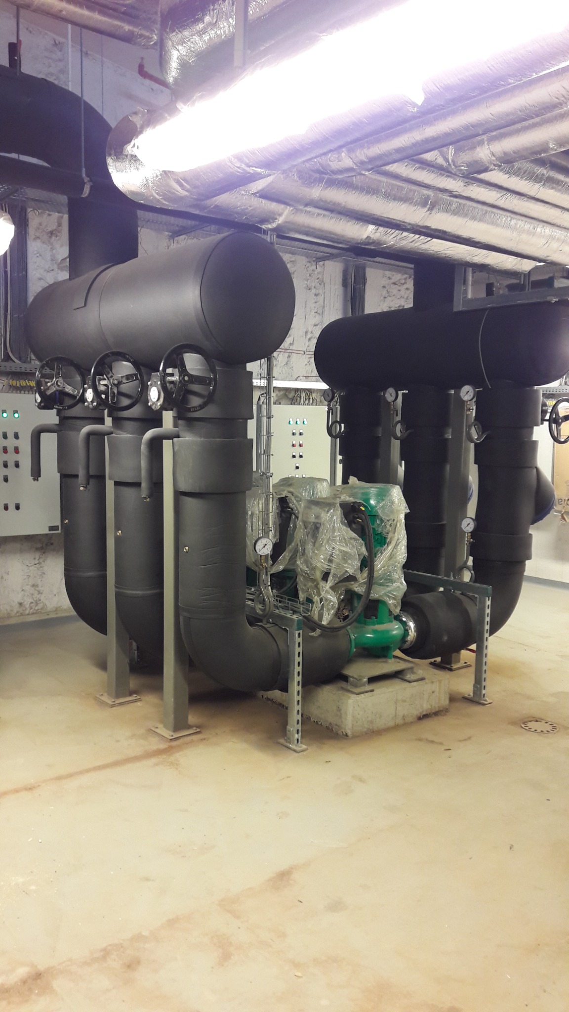 Instalacja przemysłowa z czarnych, izolowanych rur i zaworów, z zielonymi pompami zabezpieczonymi folią, na betonowej podstawie w pomieszczeniu technicznym z widocznymi przewodami wentylacyjnymi...