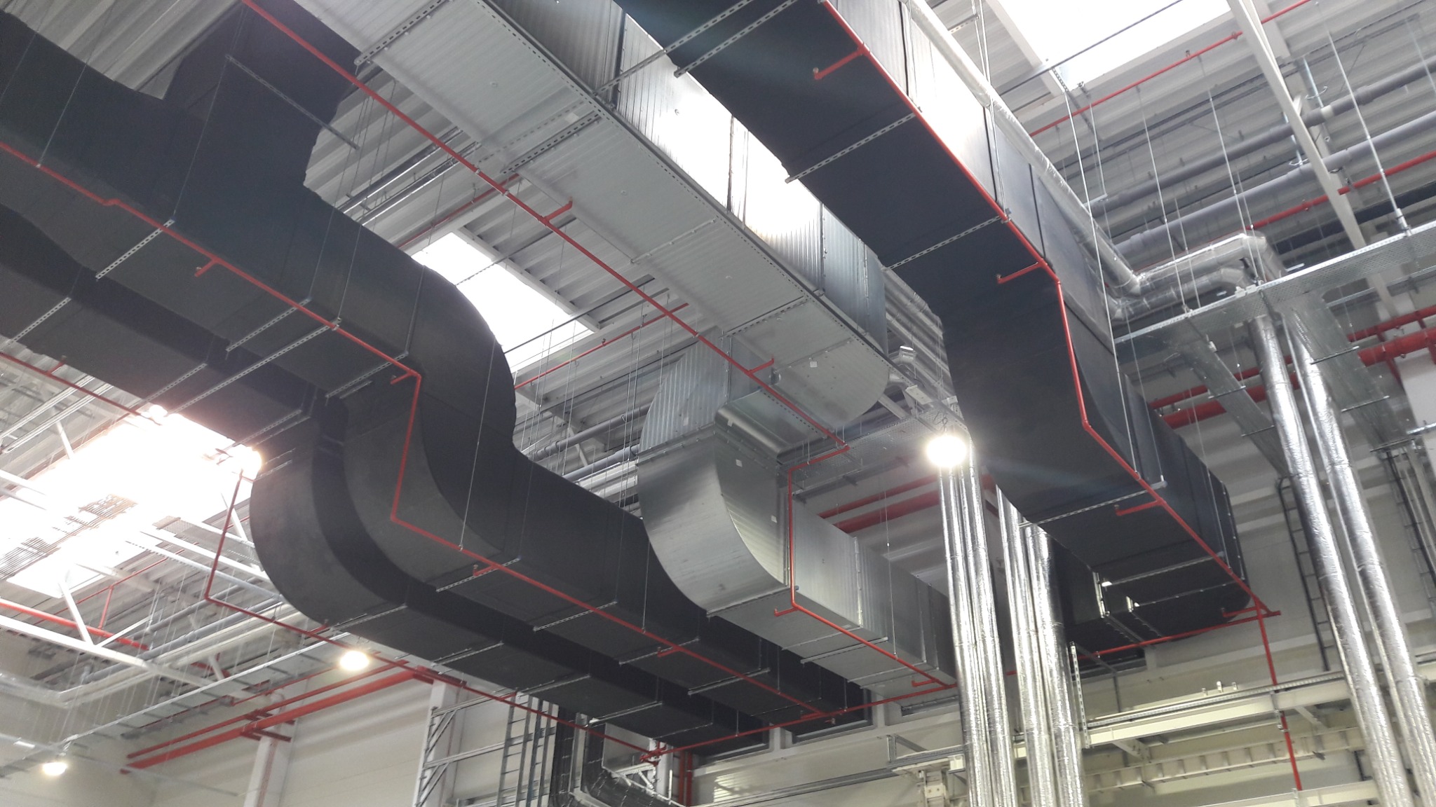 Rozbudowany system wentylacyjny z czarnymi, prostokątnymi kanałami i czerwonymi rurami w jasnym, przemysłowym wnętrzu z oknami dachowymi.