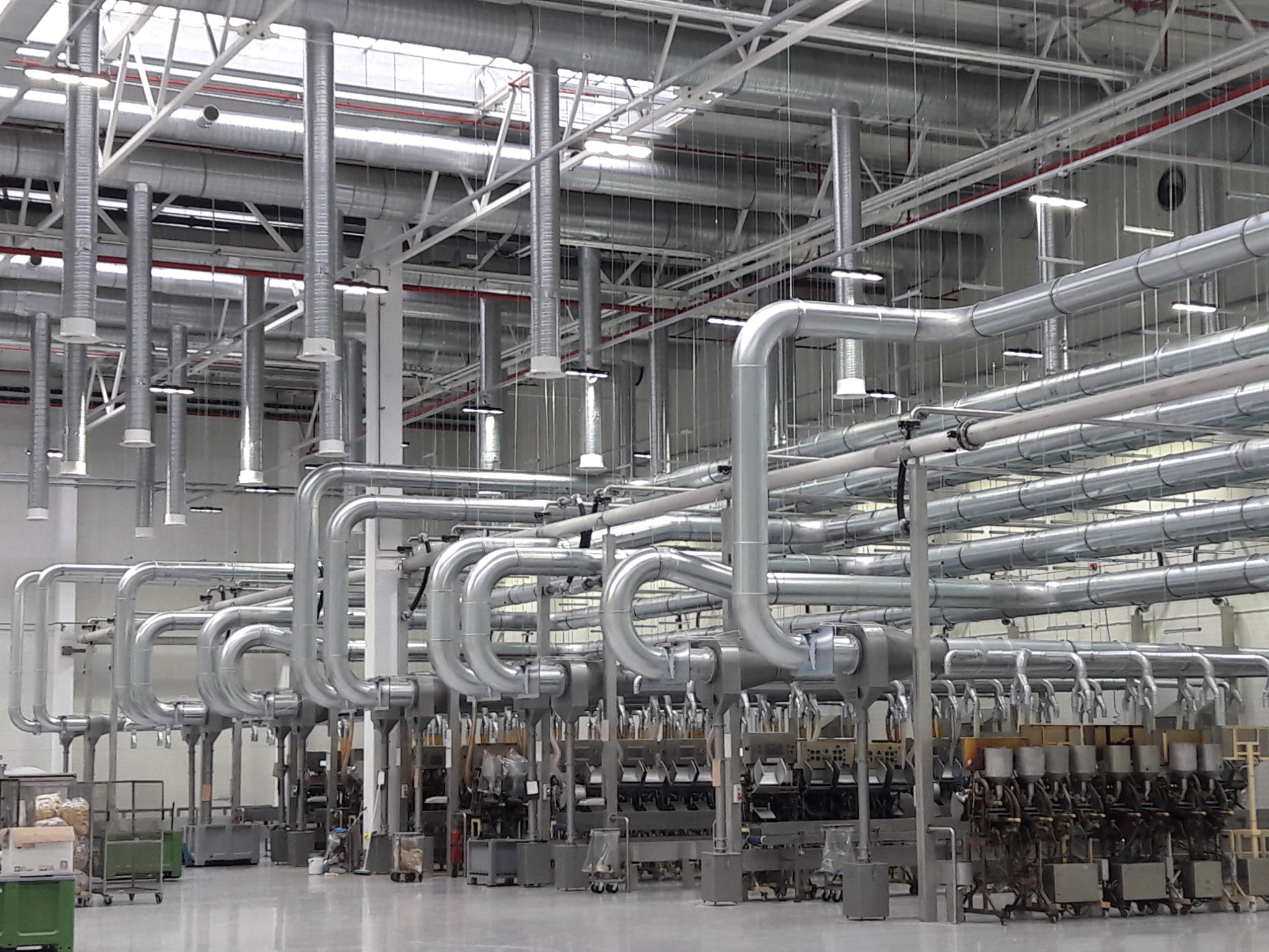Rozbudowany system rur wentylacyjnych ze stali nierdzewnej w hali produkcyjnej, widoczne liczne pionowe i poziome odcinki oraz podwieszane oświetlenie.