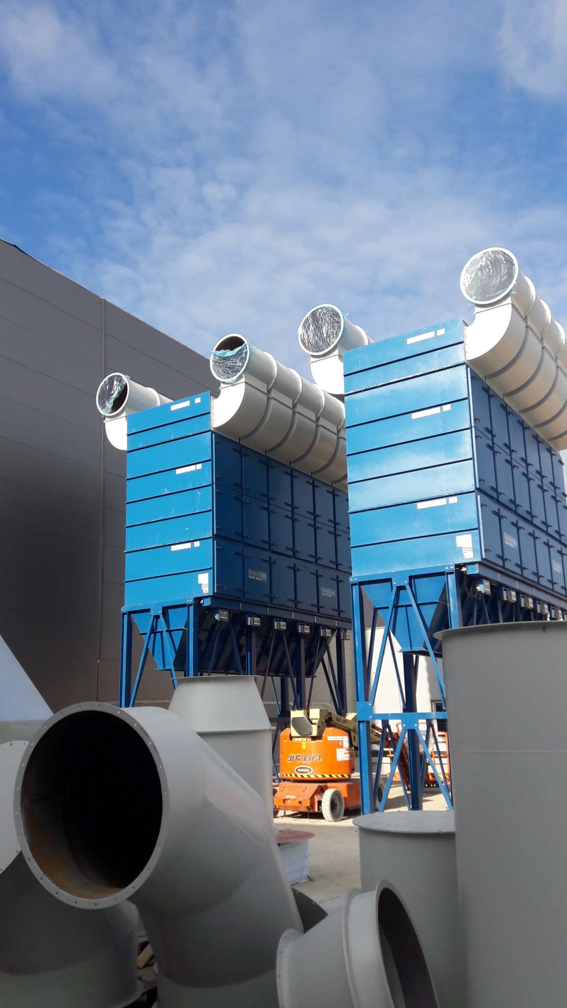 Dwa niebieskie systemy filtracji powietrza z rurami wentylacyjnymi na dachu, pomarańczowy podnośnik nożycowy w tle, widok z dołu na rury wentylacyjne na pierwszym planie.