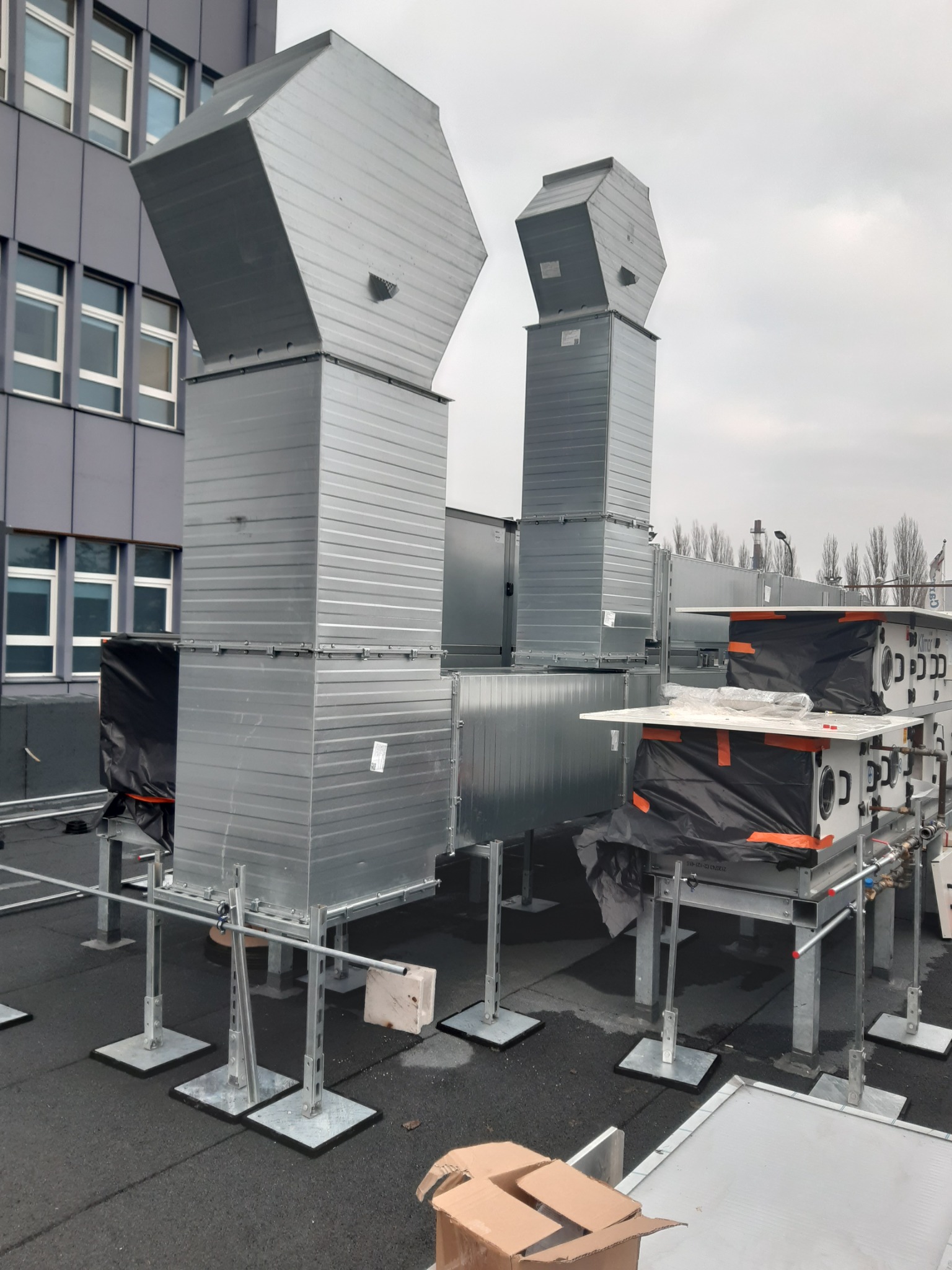 System kanałów wentylacyjnych z ocynkowanej blachy na dachu budynku komercyjnego, widoczne elementy montażowe i podesty pod rekuperatory, w tle budynek biurowy.