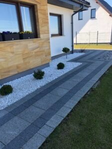 Ścieżka z szarej kostki brukowej z ciemniejszymi pasami, obłożona białym grysem, prowadząca wzdłuż elewacji domu wykończonej piaskowcem i białym tynkiem. W tle widoczny trawnik i fragment drugiego...