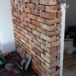 W trakcie układania dekoracyjnej ściany z cegieł w Płocku, widoczne krzyżyki dystansowe, narzędzia murarskie i odsłonięte gniazdka elektryczne.