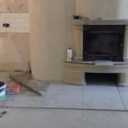 Wnętrze w trakcie remontu: ułożone płytki podłogowe, częściowo wykończona ściana z białymi płytkami, obudowany kominek z półką, widoczne puszki z farbą i narzędzia.