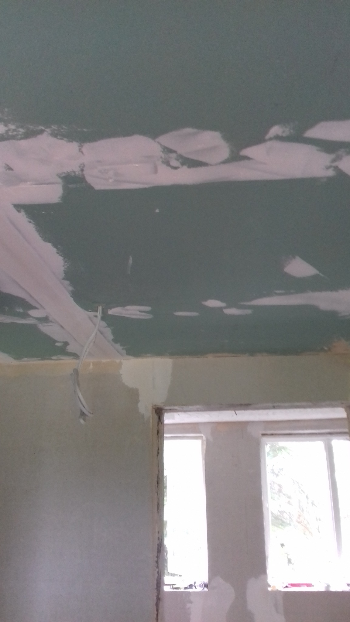 Niedokończony sufit z płyt kartonowo-gipsowych, widoczne łączenia zaszpachlowane, zwisające kable, ściany przygotowane do malowania, okno z widokiem na zieleń.