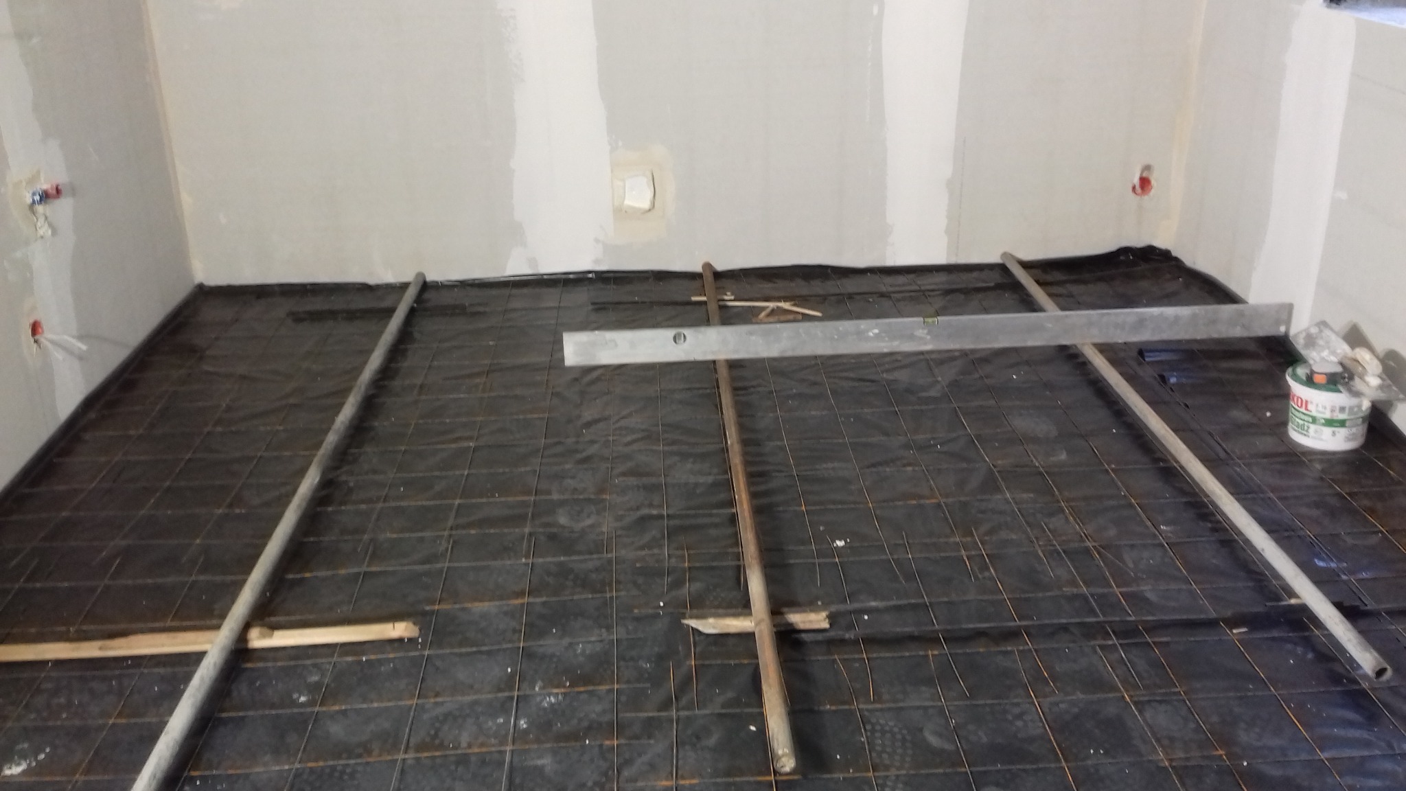 Przygotowanie podłoża pod ogrzewanie podłogowe w łazience w Płocku: czarna folia, metalowa siatka, poziomica i rury na tle surowych ścian.