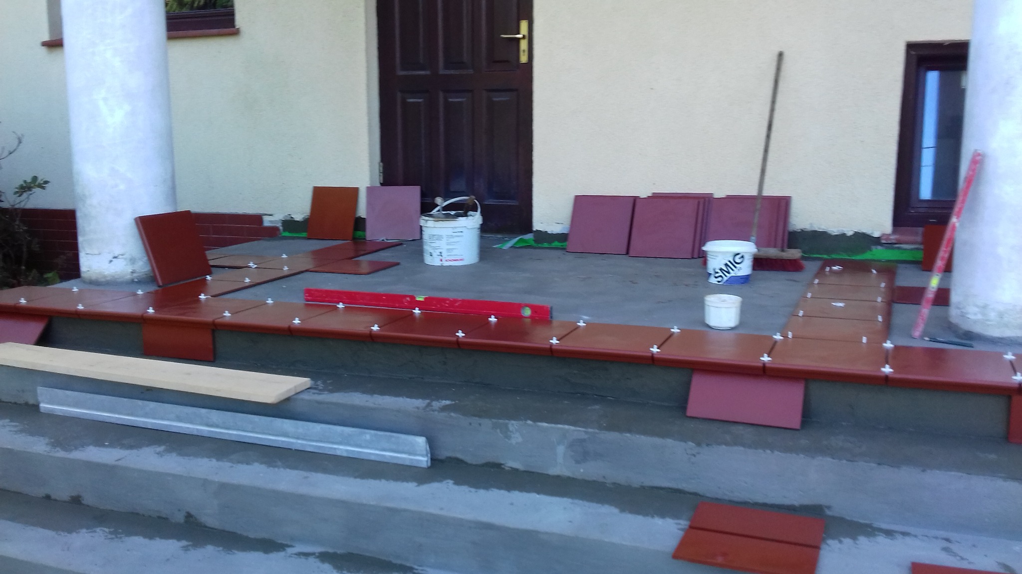 Brązowe płytki kładzione na schodach wejściowych do domu, widoczny poziomica, wiadro z klejem i krzyżyki dystansowe.