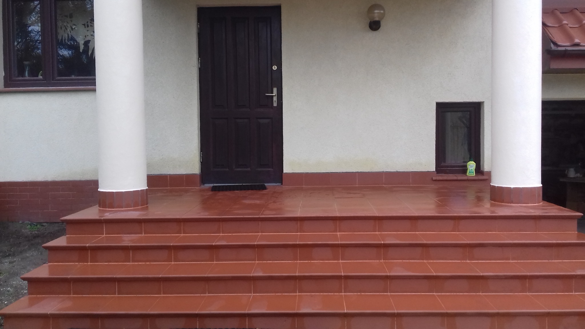 Kafelkowane schody wejściowe w odcieniach terakoty prowadzące do ciemnych drzwi wejściowych, z kolumnami i małym oknem po bokach, całość oświetlona lampą zewnętrzną.