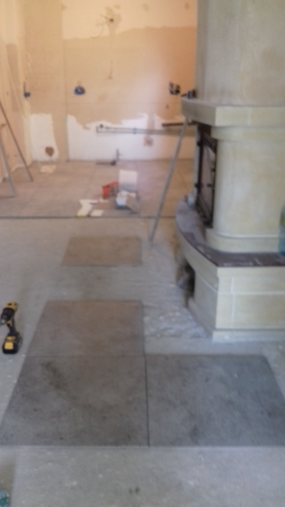 Fragment remontowanego pomieszczenia z położonymi częściowo szarymi płytkami podłogowymi, widoczne narzędzia i elementy przygotowania do prac wykończeniowych przy ścianach oraz kominku.