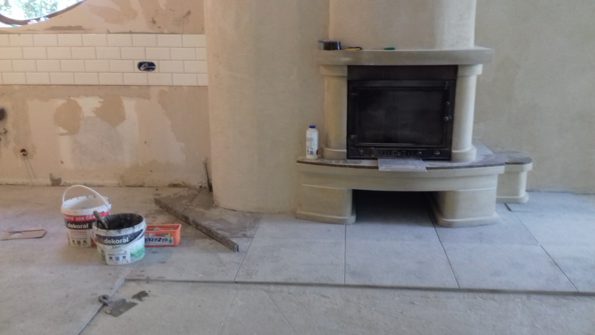 Wnętrze w trakcie remontu: ułożone płytki podłogowe, częściowo wykończona ściana z białymi płytkami, obudowany kominek z półką, widoczne puszki z farbą i narzędzia.