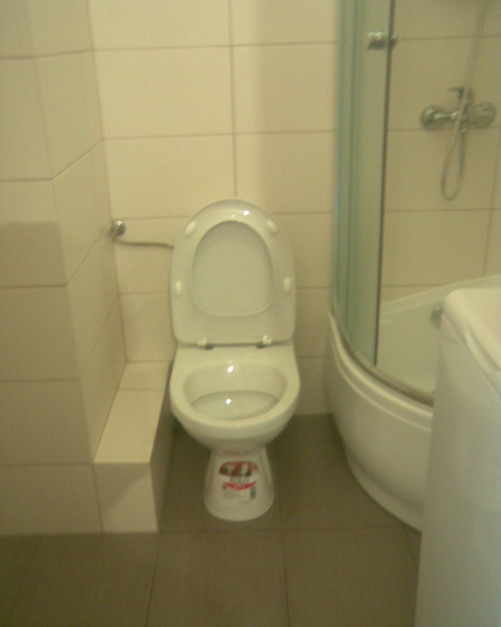 Standardowa toaleta z otwartą klapą, obok kabina prysznicowa z półokrągłą podstawą, widoczne szare płytki podłogowe i beżowe płytki ścienne.