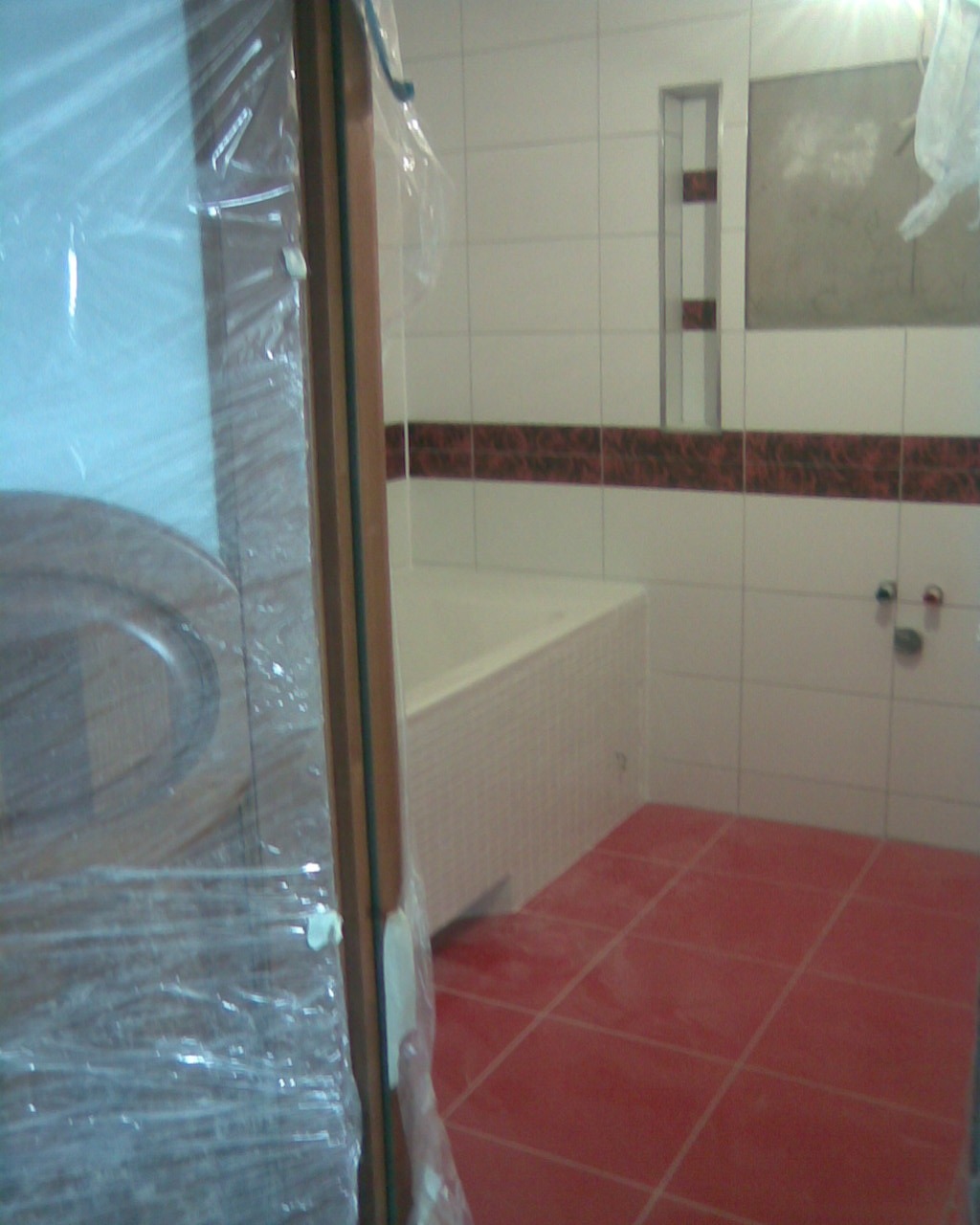 Nowa łazienka z czerwonymi płytkami podłogowymi i białą wanną, widok przez zapakowane w folię drzwi, ściany wyłożone białymi płytkami z bordiurą.