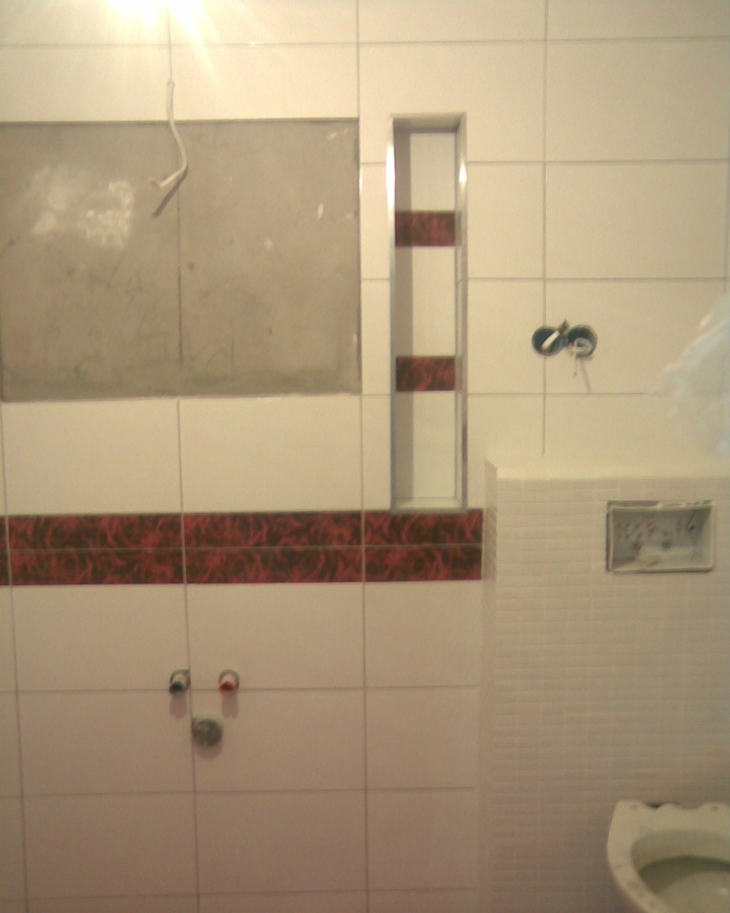 Niedokończony remont łazienki: białe płytki z bordowym pasem dekoracyjnym, wnęka w ścianie z półkami, przygotowane przyłącza wodne i kanalizacyjne, częściowo zamontowana zabudowa WC.