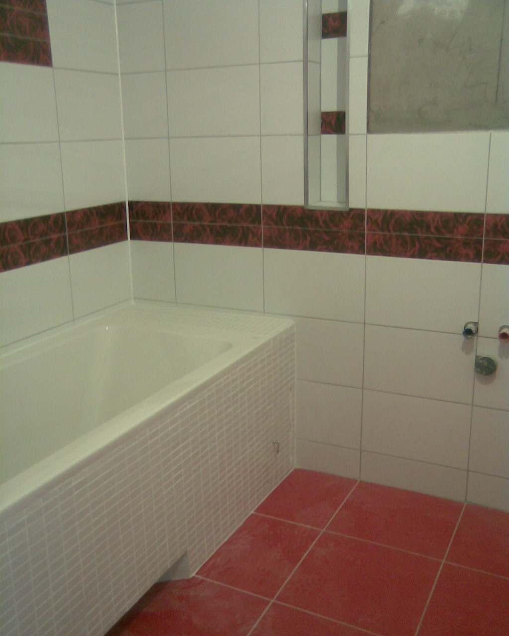 Wykończona łazienka z biało-czerwonymi płytkami, widoczna wanna obudowana mozaiką, czerwona podłoga i wnęka w ścianie.