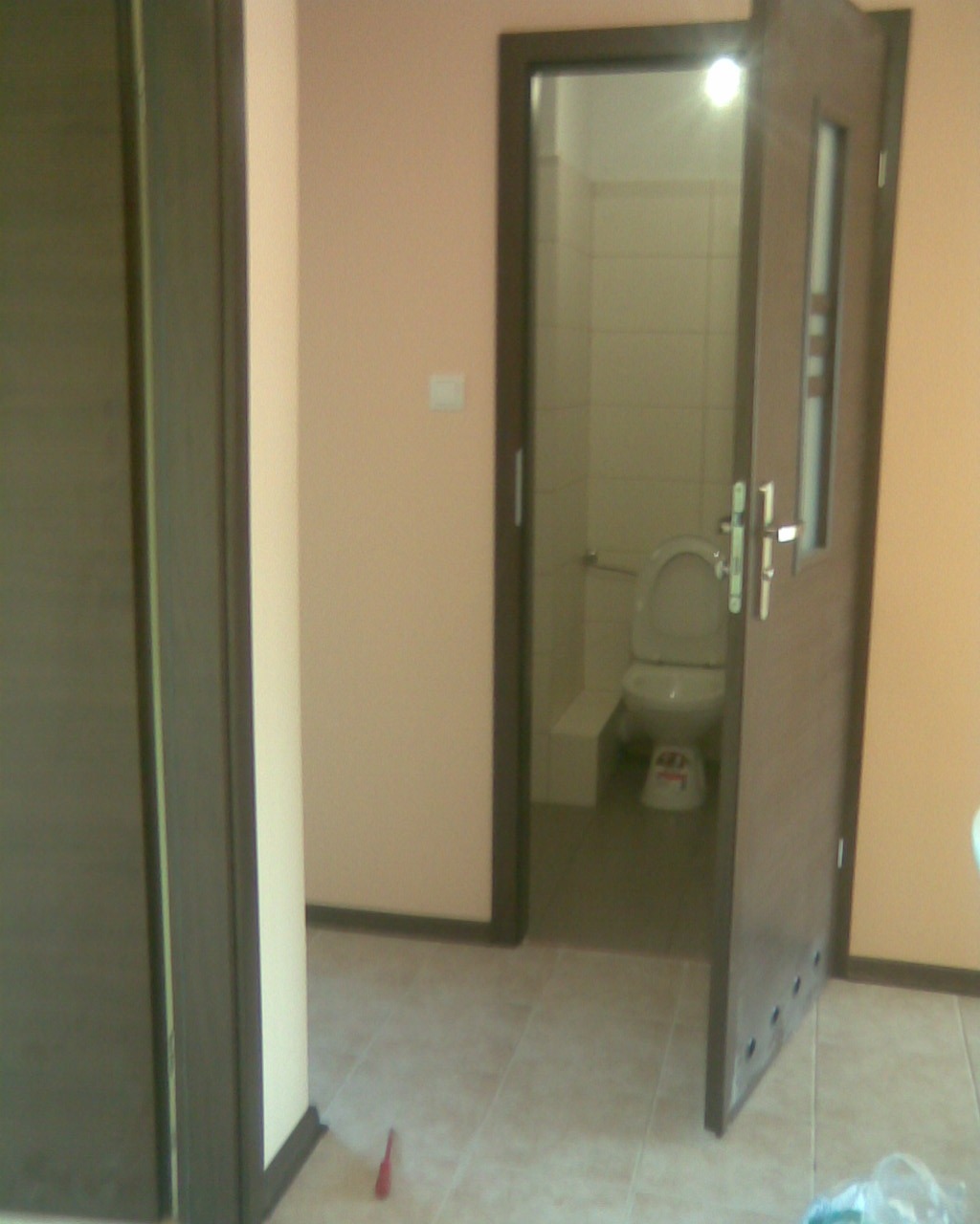 Otwarta, ciemnobrązowa ościeżnica z szybką, ukazująca wnętrze wyremontowanej łazienki z toaletą w Płocku. Na podłodze widoczny fragment narzędzia i folii ochronnej.