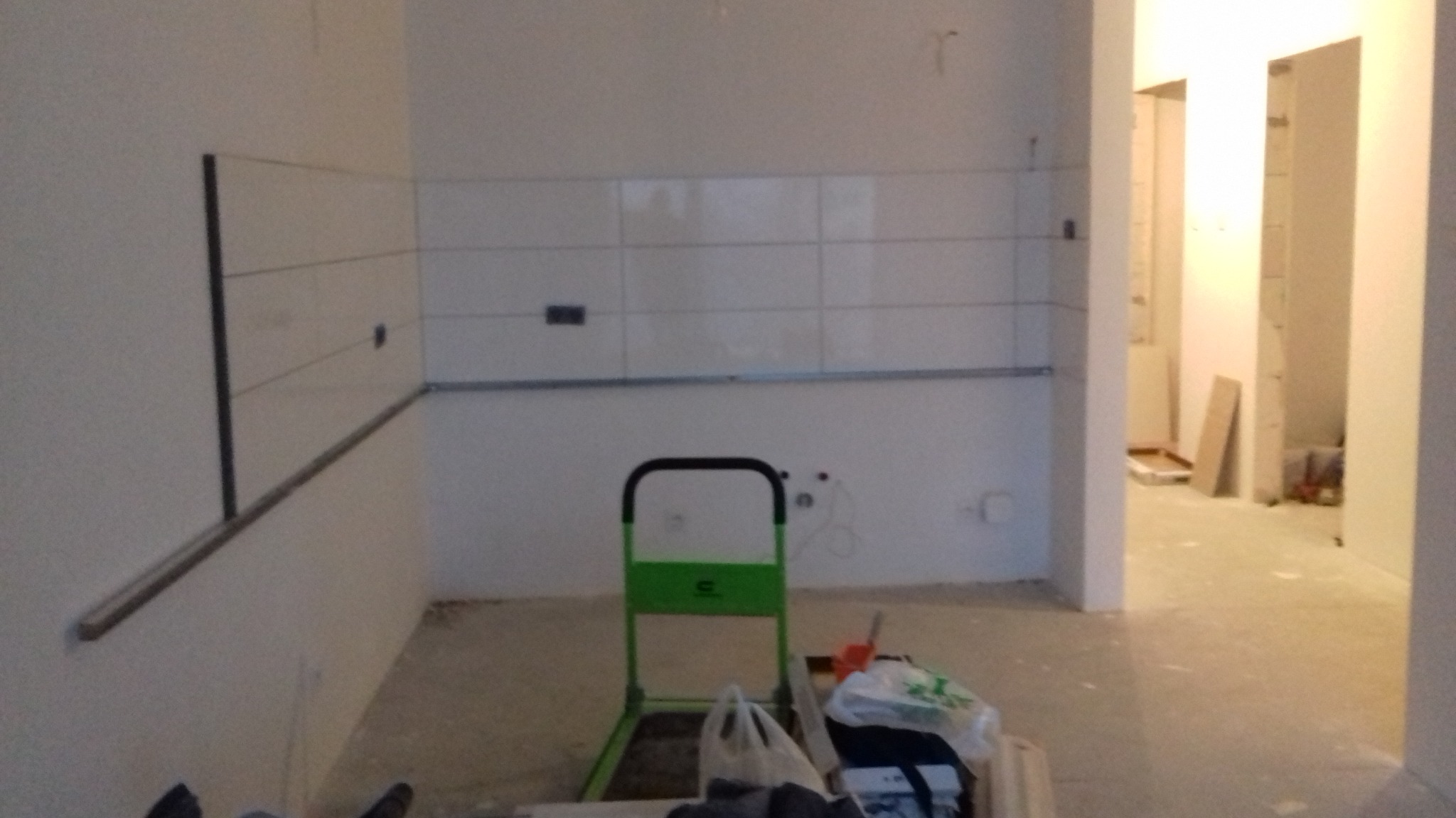 Wnętrze w trakcie wykańczania: białe płytki ułożone na ścianie, widoczne przewody elektryczne, zielona drabinka i torby z materiałami budowlanymi na podłodze, perspektywa na sąsiednie pomieszczenie.