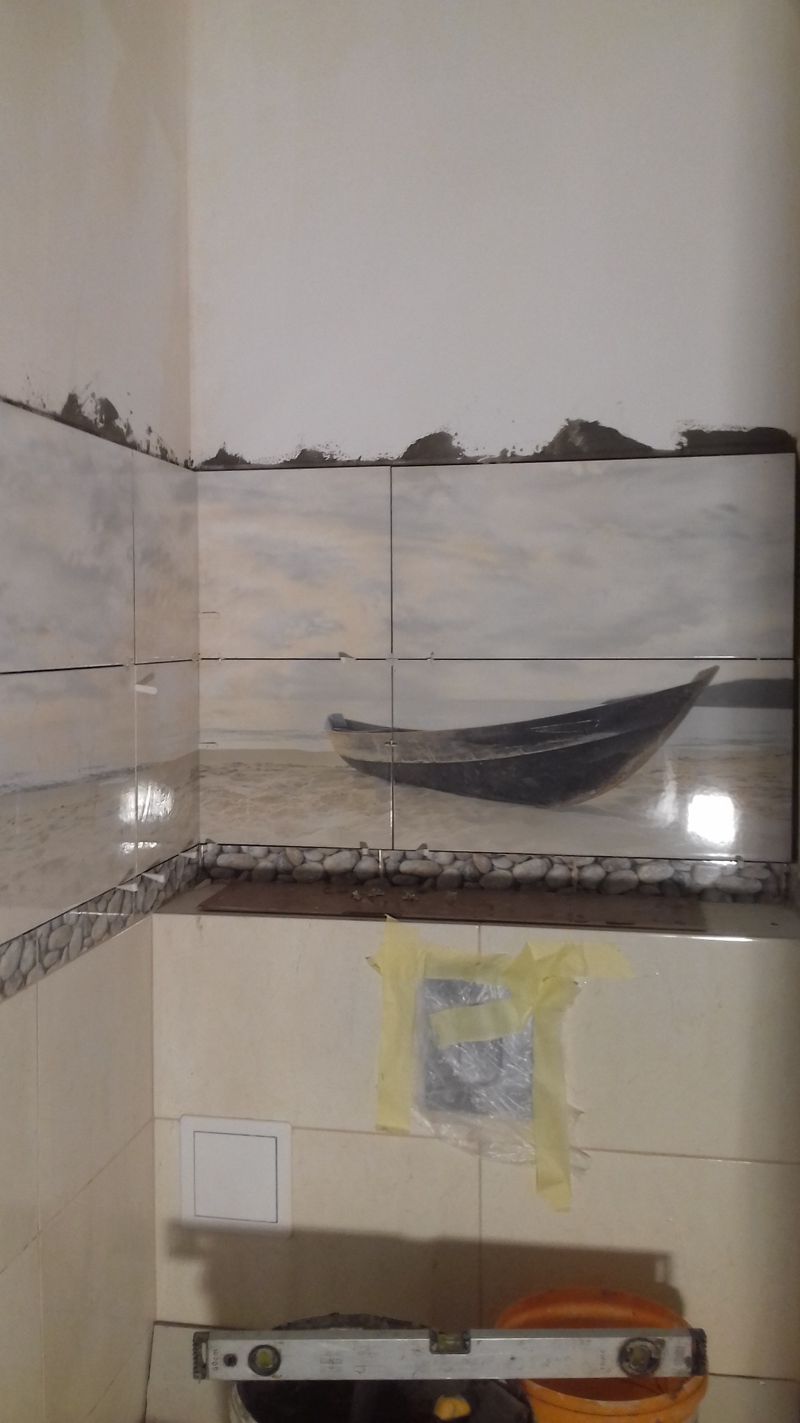 Fragment ściany w trakcie wykańczania z ułożonymi płytkami z motywem łodzi na plaży, widoczne narzędzia i zabezpieczone elementy instalacji.