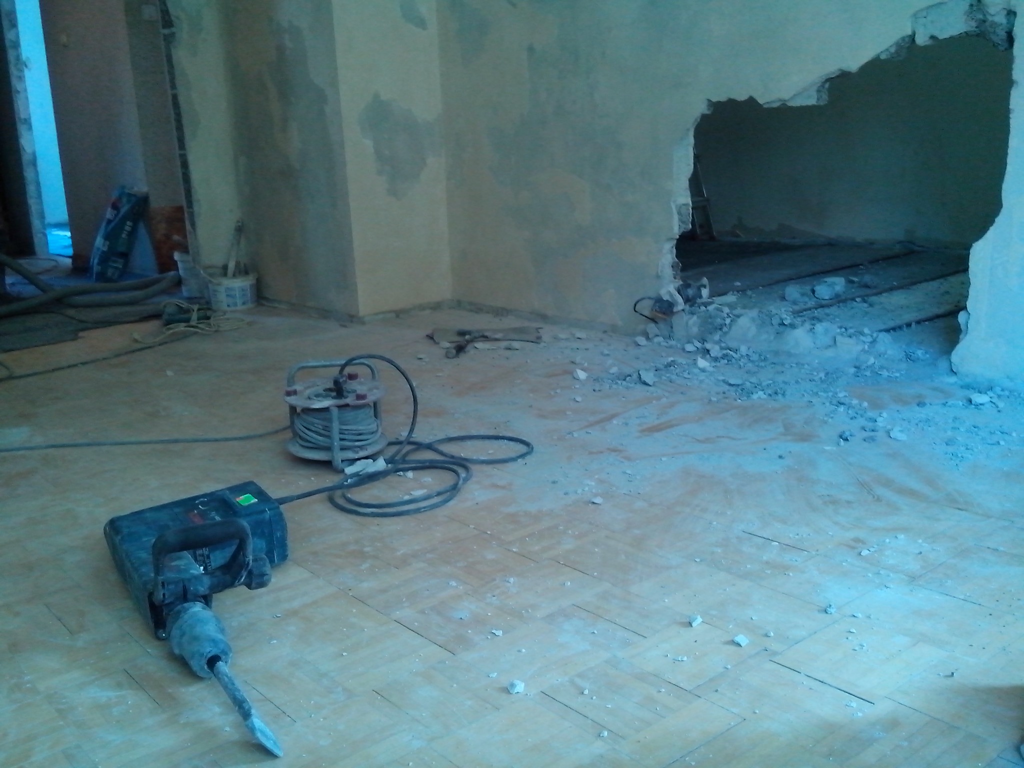 Wnętrze w trakcie prac demontażowych: wyburzona część ściany z widocznymi deskami, na podłodze leży młot udarowy z przewodem zwiniętym na szpuli, gruz i pył na podłodze z parkietu.