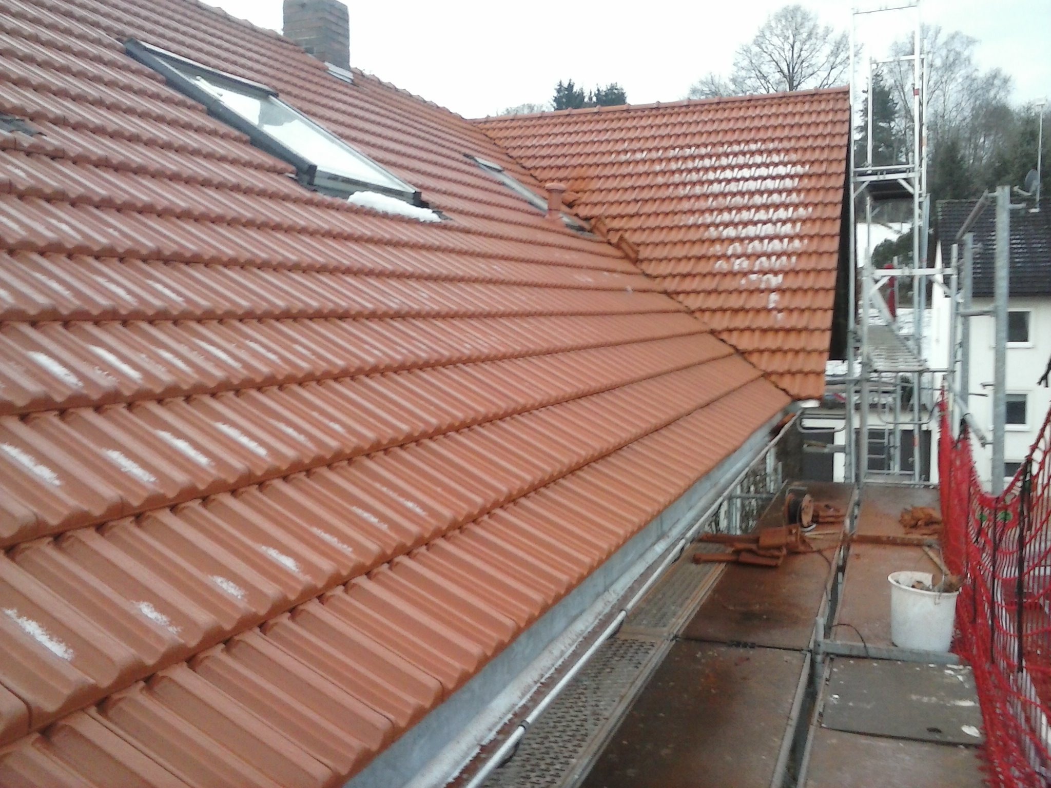 Czerwona dachówka ceramiczna na skośnym dachu z oknem dachowym, częściowo pokryta śniegiem, widok z rusztowania, w tle drzewa i budynek.