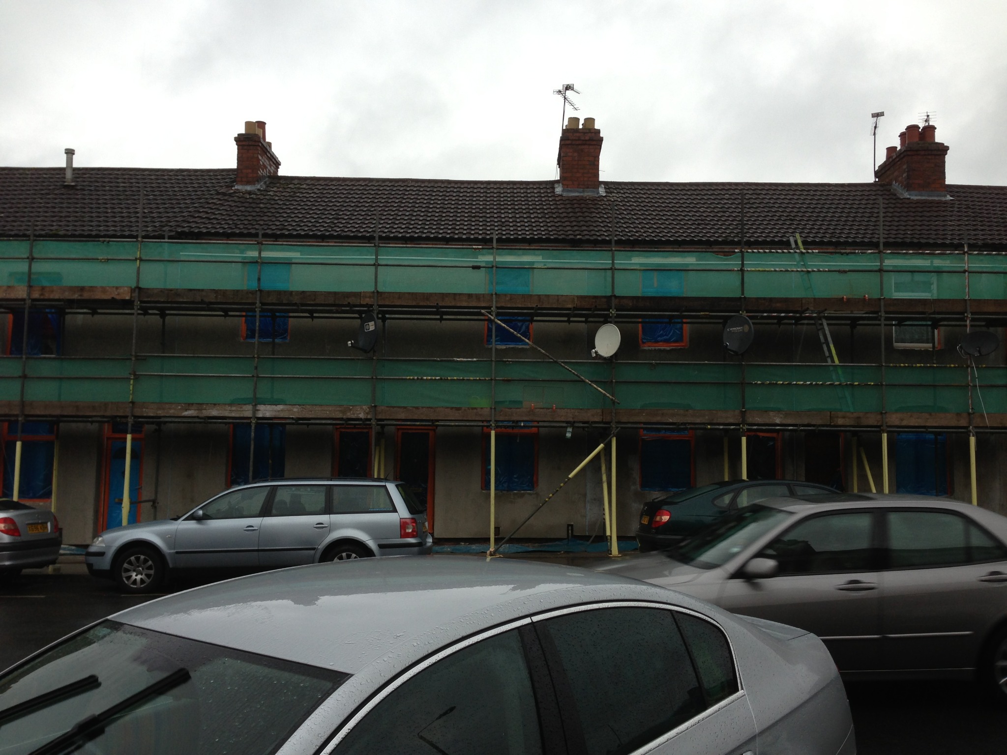 Budynek mieszkalny w trakcie remontu elewacji, osłonięty rusztowaniami i siatką ochronną, z zaparkowanymi samochodami na pierwszym planie w pochmurny dzień.