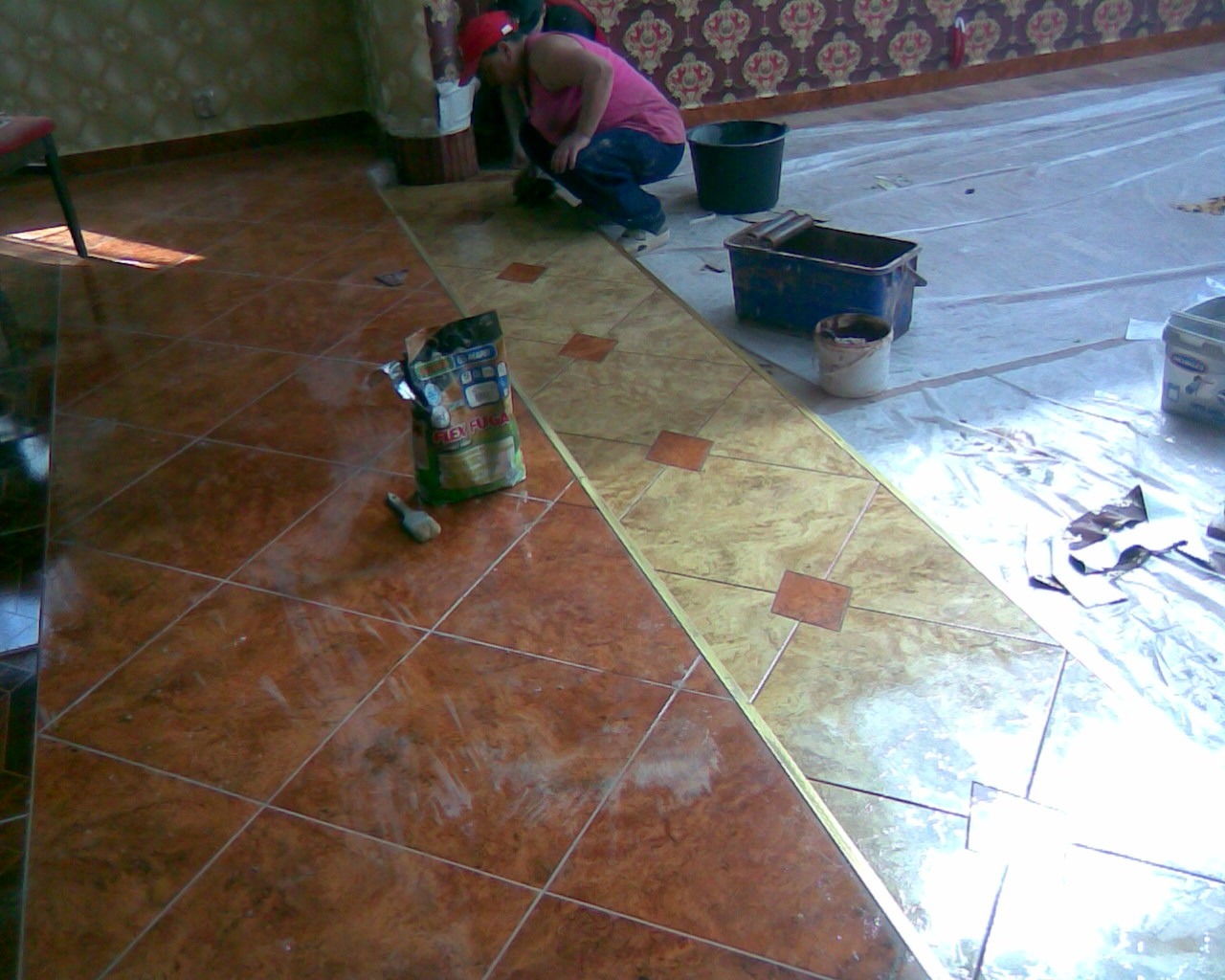 Układanie płytek podłogowych w Płocku: mężczyzna w czerwonej czapce fuguje nowo położoną podłogę z płytek w dwóch odcieniach brązu, z widocznym workiem fugi i folią ochronną na części podłogi.