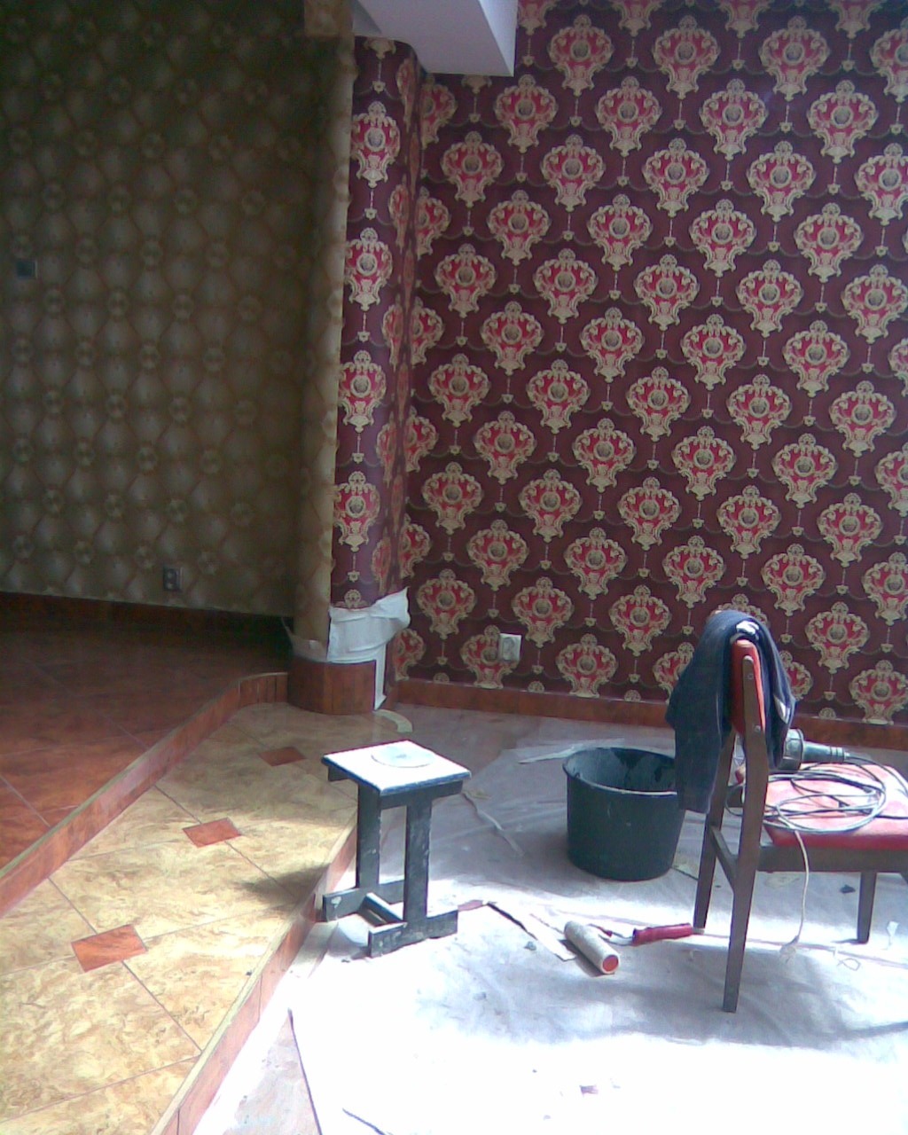 Pokój w trakcie tapetowania; widoczna część ściany z nową bordową tapetą w ornamenty, fragment ściany z odklejoną starą tapetą, oraz narzędzia: wiadro, wałek, stołek i krzesło z narzuconą kurtką.