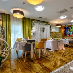 Uroczysta sala bankietowa z dekoracjami kwiatowymi, białymi obrusami, eleganckimi krzesłami z kokardami, podświetlanym napisem oraz kolorowym obrazem na ścianie.