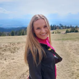 Uśmiechnięta blondynka w czarno-różowej kurtce i z paskiem Guess na tle górskiego krajobrazu z drewnianymi domkami.