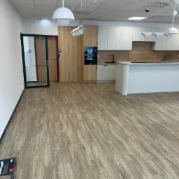 Jasne panele podłogowe w nowym biurze z aneksem kuchennym. Widoczne narzędzia do montażu paneli w lewym dolnym rogu.