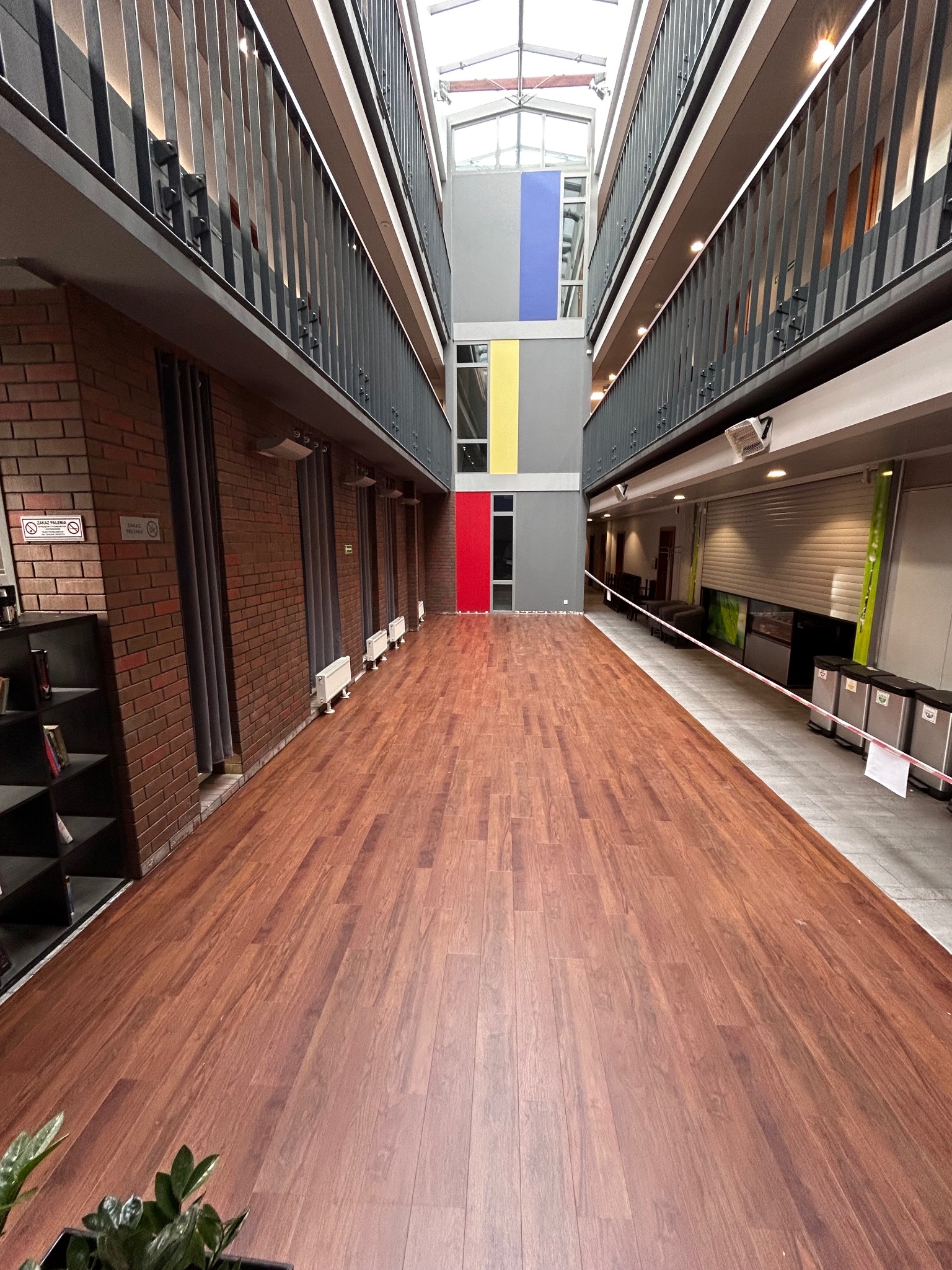 Nowa, brązowa podłoga z paneli ułożona w korytarzu biurowca z przeszklonym dachem i balkonami po bokach. Widoczne grzejniki i zasłony wzdłuż jednej ściany.