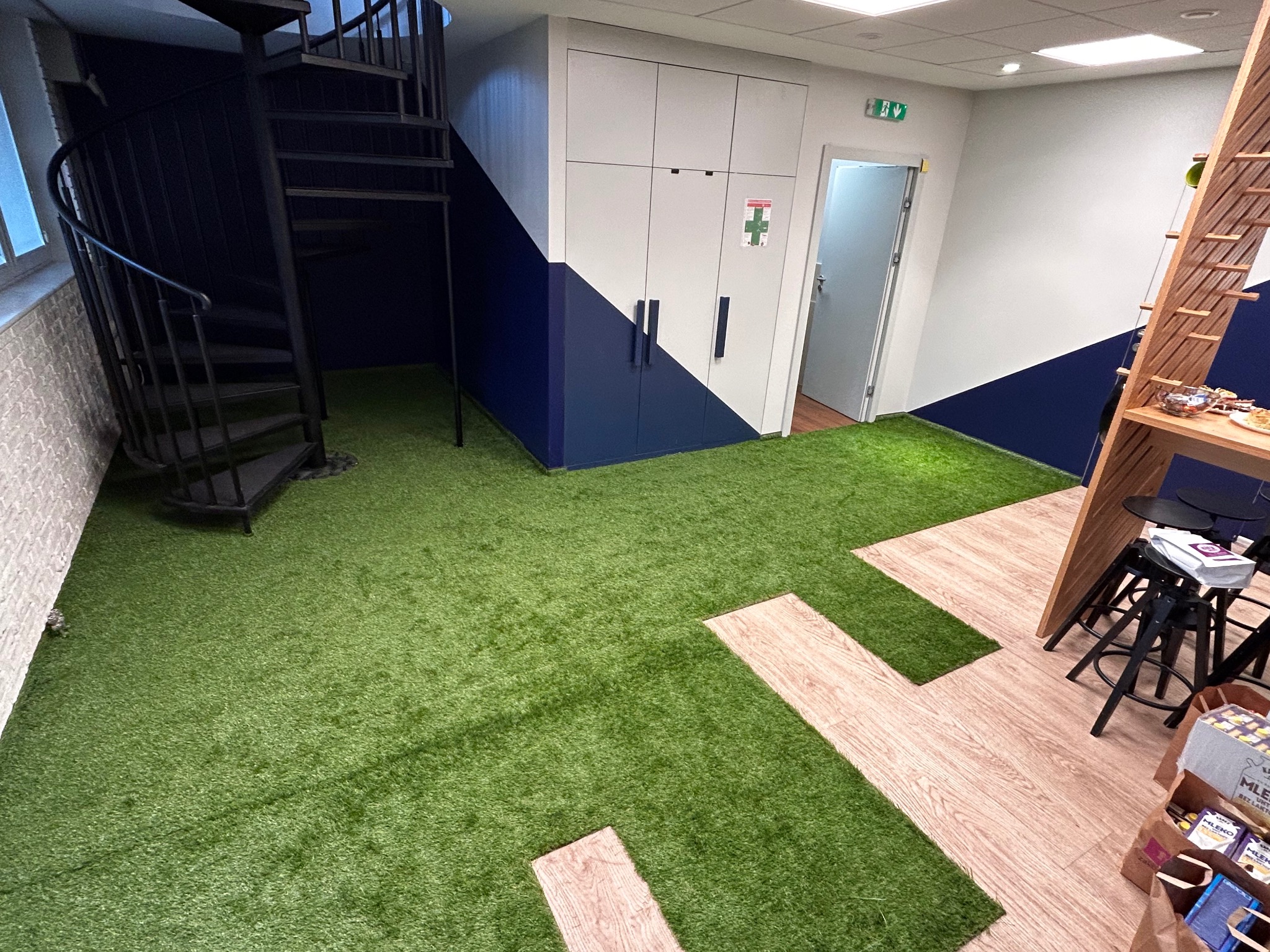 Wnętrze biura z zieloną wykładziną imitującą trawę, fragmentami paneli podłogowych, spiralnymi schodami i zabudową meblową w kolorach bieli i granatu.