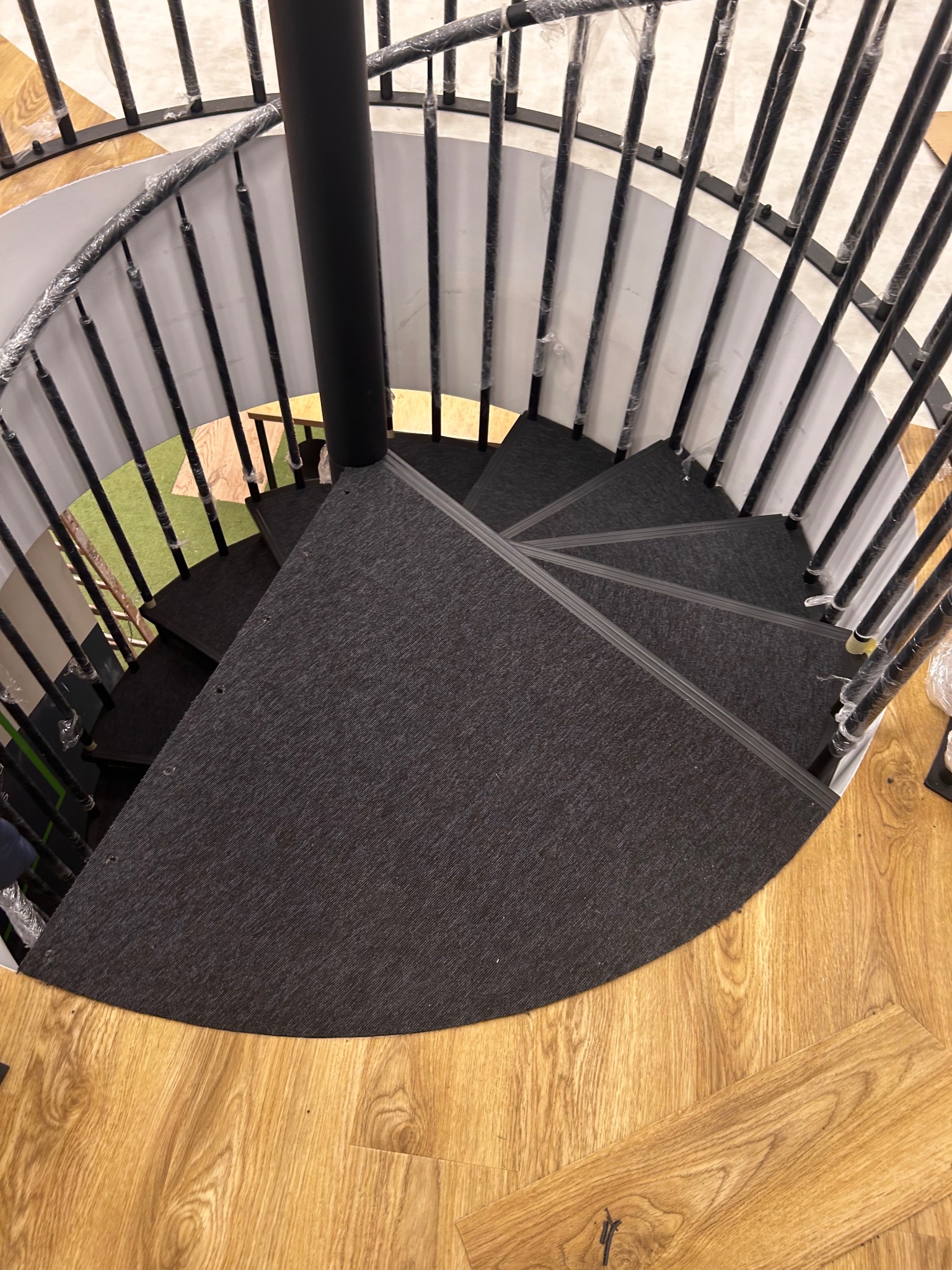 Widok z góry na spiralne schody z czarną metalową balustradą, stopnie pokryte ciemną wykładziną dywanową, a podłoga wykończona panelami imitującymi drewno.