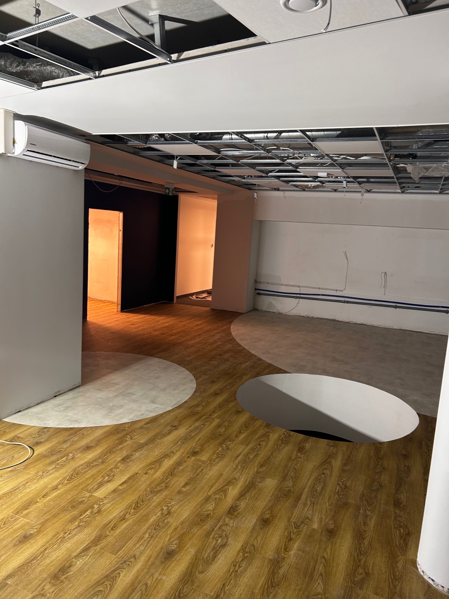 Wnętrze w trakcie remontu z drewnopodobną podłogą z wyciętymi okręgami, widoczne instalacje pod sufitem i klimatyzator.