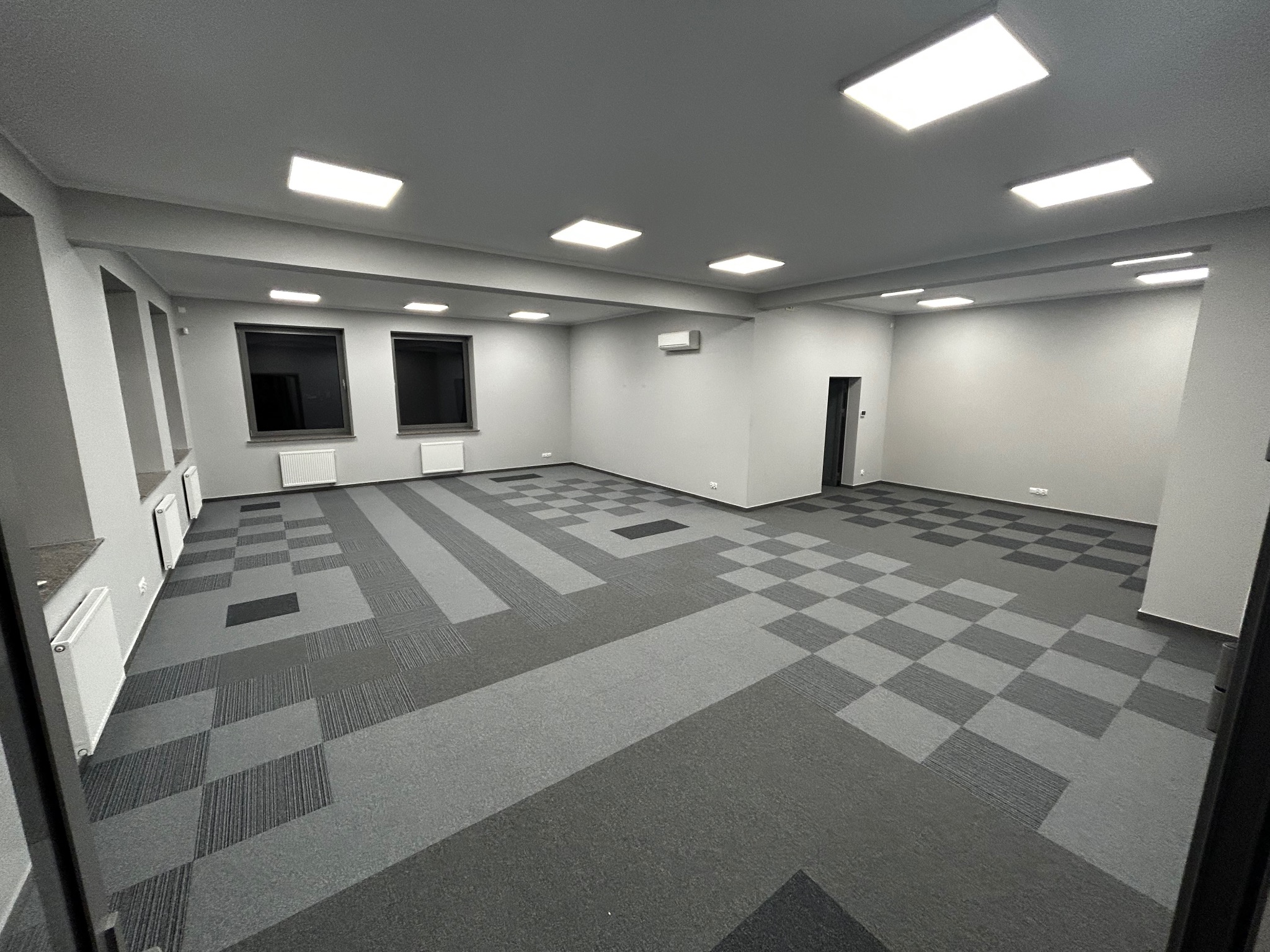 Nowoczesna, pusta przestrzeń biurowa z szarą wykładziną dywanową ułożoną w geometryczne wzory, widoczne okna z grzejnikami i równomiernie rozmieszczone oświetlenie sufitowe.