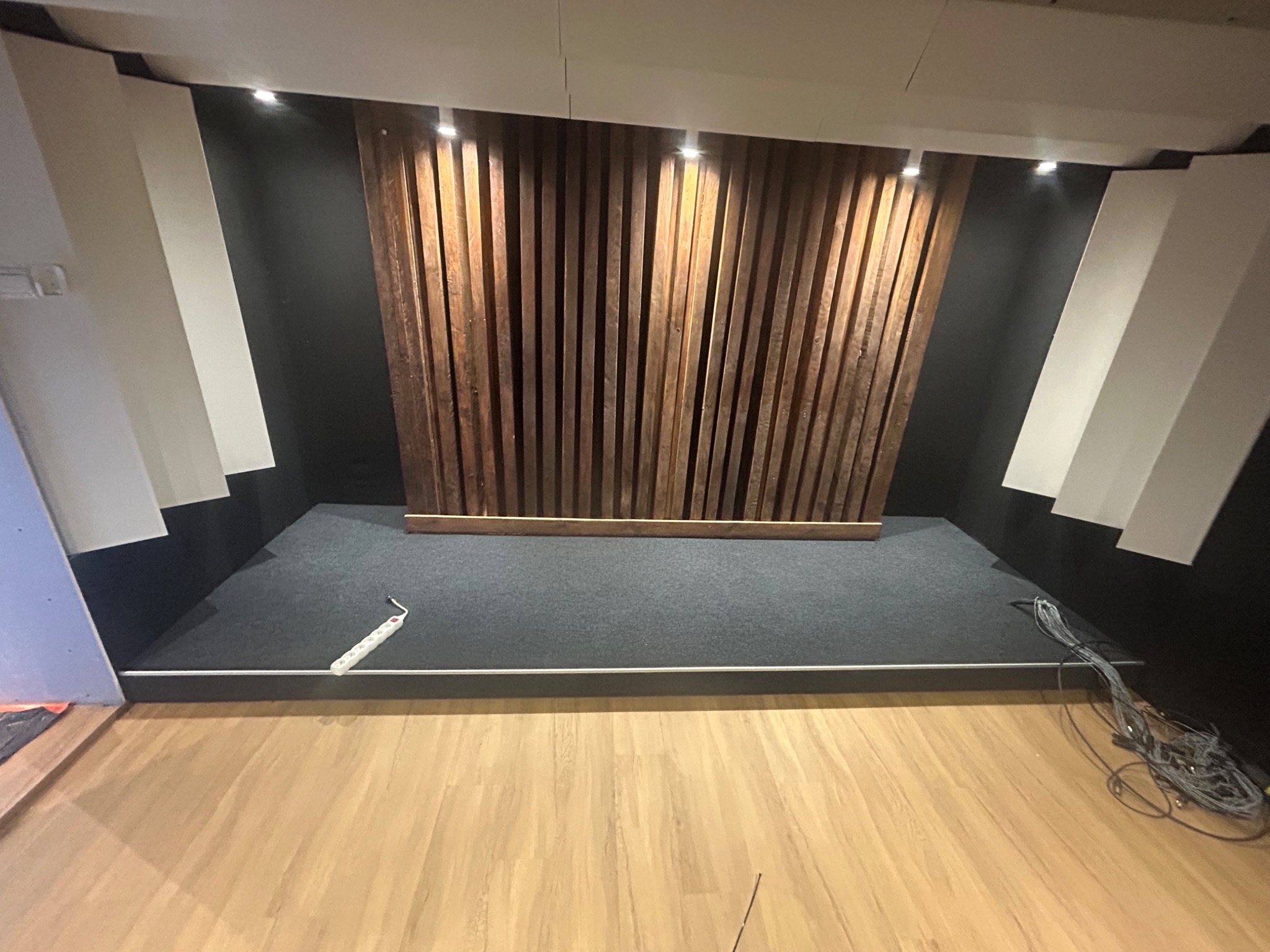 Wykończone wnętrze studia nagraniowego z panelami akustycznymi z drewna na ścianie, szarą wykładziną podłogową i drewnianą podłogą, widoczna listwa przypodłogowa i przewody.